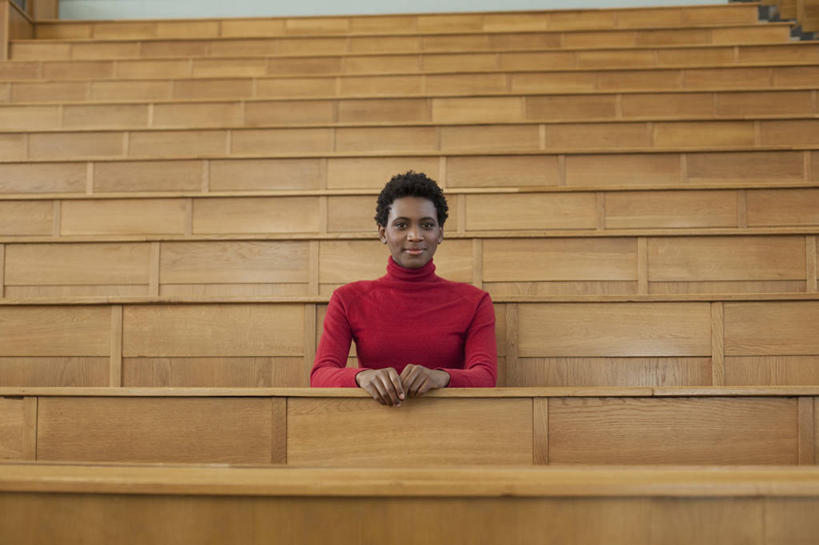 西方人,一个人,学校,教学楼,坐,笑,微笑,横图,室内,白天,正面,毛衣,桌子,欧洲,非洲,黑人,欧洲人,仅一个年轻女性,仅一个女性,仅一个人,木制,墙,注视,书桌,校园,墙壁,墙面,红色,课桌,木制品,观察,看,教室,观看,察看,关注,线衫,毛线衣,绒线衣,年轻女性,女人,女性,非洲人,坐着,半身,彩图,绒衣,套衫,套头毛衣,线衣,教学大楼,报告厅