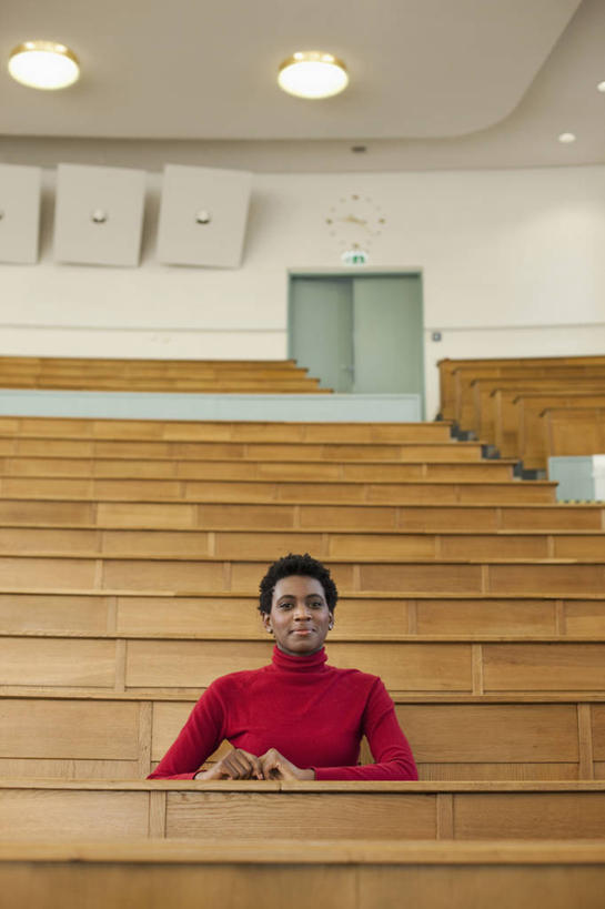 西方人,一个人,学校,教学楼,坐,笑,微笑,竖图,室内,白天,正面,毛衣,桌子,照明,欧洲,非洲,黑人,欧洲人,仅一个年轻女性,仅一个女性,仅一个人,木制,墙,灯光,注视,书桌,校园,顶灯,墙壁,墙面,灯,红色,课桌,木制品,灯具,观察,看,教室,观看,照亮,察看,照明设备,关注,线衫,毛线衣,绒线衣,年轻女性,女人,女性,非洲人,坐着,半身,彩图,绒衣,套衫,套头毛衣,线衣,教学大楼,报告厅